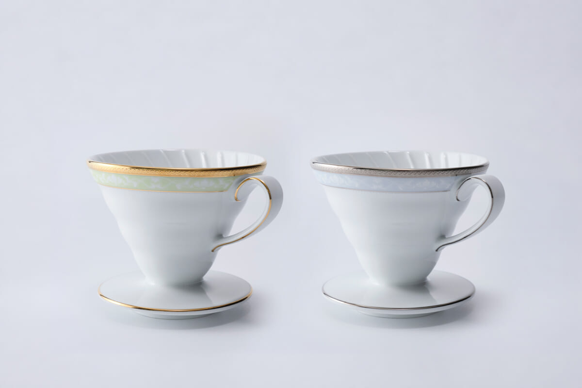 Two white porcelain teacups with matching saucers side by side on a pale background one with gold trim and a pale green accent and the other with silver trim