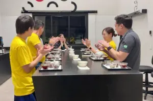 00e852dbdd6b1c36d8c84822c7724822 | コーヒーステーション Six people around a long black table in a bright room clapping after a tasting with small bowls and cups on trays