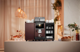 Professional espresso machine on a kitchen counter with cups on top and an open mug rack beside it in warm ambient light