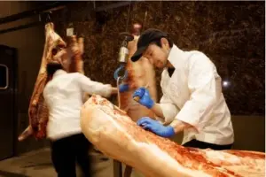 Two butchers carve a large leg of meat on a processing table one wears a cap and blue gloves while trimming