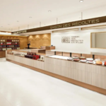 Modern bakery interior with a long pastry counter displaying cakes and shelves of products beneath a warm lighted ceiling SAISON DES RÉCOLTES sign above