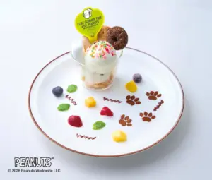 Glass of ice cream in a small cup topped with colorful sprinkles and a round cookie set on a white plate with decorative sauces fruits and paw print chocolate shapes Peanuts branding visible in corner