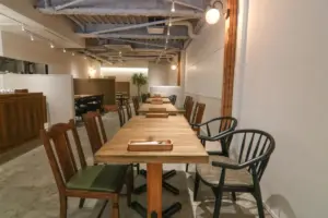 Long wooden communal table with mixed wooden and black chairs in an industrial style cafe exposed ductwork and warm lighting