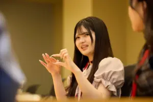 Woman with long dark hair gestures with her hands while speaking in a meeting room wearing a light blouse and a red lanyard label