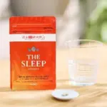 Red pouch labeled THE SLEEP with a few white pills on a small plate beside a glass of water on a wooden table