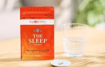Red pouch labeled 'THE SLEEP' with a few white pills on a small plate beside a glass of water on a wooden table.