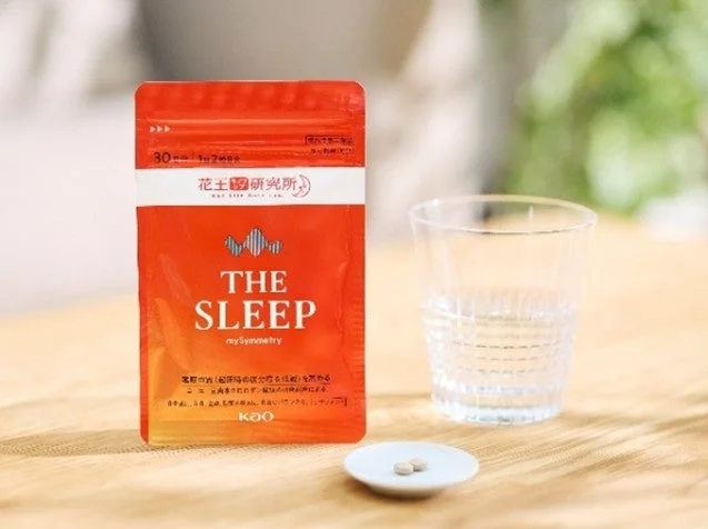Red pouch labeled THE SLEEP with a few white pills on a small plate beside a glass of water on a wooden table