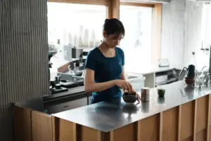 Barista in a blue shirt preparing coffee at a stainless steel counter in a bright cafe