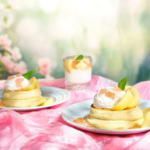 Two stacks of pancakes topped with whipped cream and peach slices on a pink satin tablecloth with a mint garnish