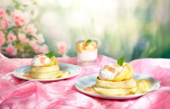 Two stacks of pancakes topped with whipped cream and peach slices on a pink satin tablecloth with a mint garnish