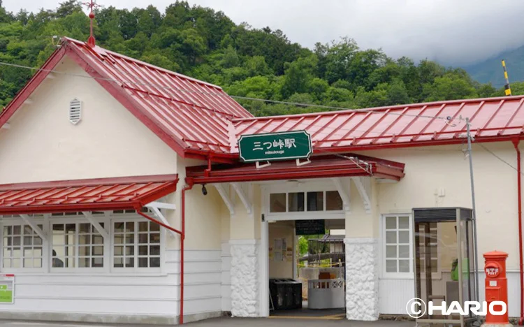 Small train station with white walls and a red roof green Mitsutoge Station sign set against green hills