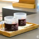 Two glass coffee mugs with dark beverage sit on a wooden serving tray Hario logo visible in a cafe setting