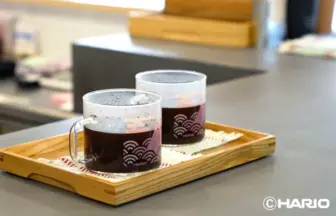 Two glass coffee mugs with dark beverage sit on a wooden serving tray Hario logo visible in a cafe setting
