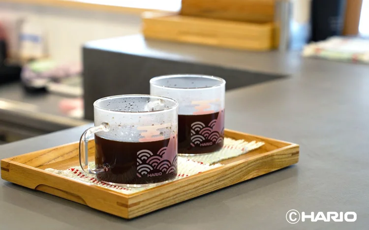 Two glass coffee mugs with dark beverage sit on a wooden serving tray Hario logo visible in a cafe setting