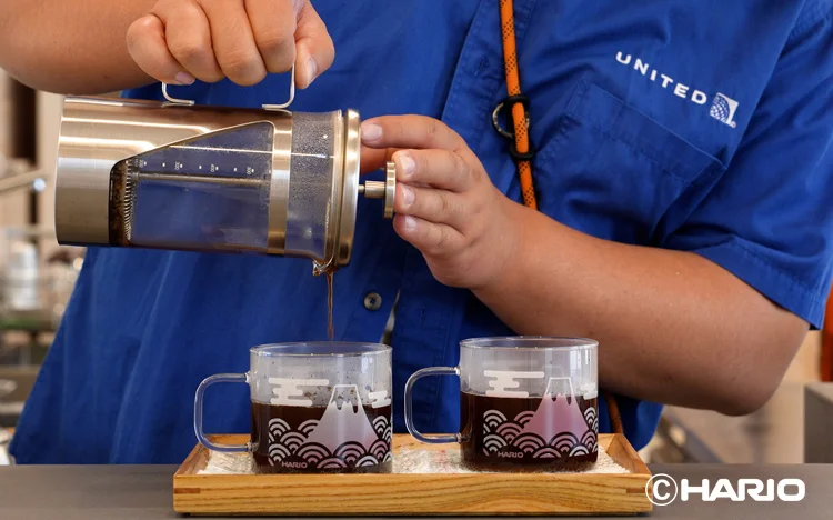 Barista in a blue uniform pours coffee from a metal siphon brewer into two glass mugs on a wooden tray