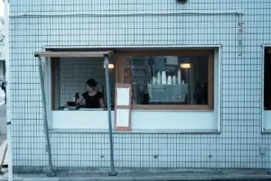 Barista serves at a small window counter in a blue tiled building coffee cups and machine visible inside