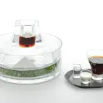 Elegant glass beverage set featuring a crystal decanter atop an ice block with herbs around the base plus a side tray with a dark drink and small cream and sugar vessels