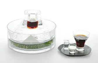 Elegant glass beverage set featuring a crystal decanter atop an ice block with herbs around the base, plus a side tray with a dark drink and small cream and sugar vessels.