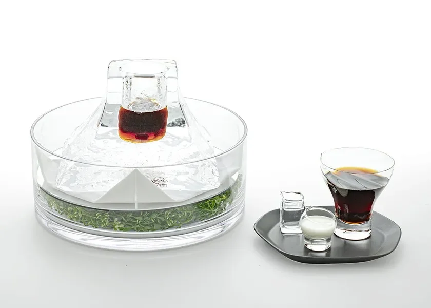 Elegant glass beverage set featuring a crystal decanter atop an ice block with herbs around the base plus a side tray with a dark drink and small cream and sugar vessels