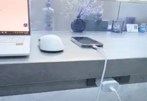A white laptop on the left a white wireless mouse and a smartphone plugged in with a white charging cable on a gray desk with decorative blue glass vases in the background
