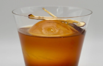 Tumbler glass of amber cocktail with a large spherical ice ball and a lemon twist garnish.