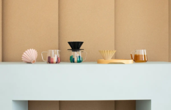 Assorted pour over coffee gear on a white shelf against a kraft paper backdrop filter cones glass carafes and a wooden stand