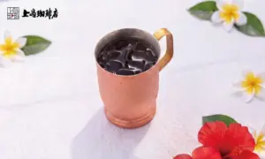Copper mug filled with iced coffee on a white table surrounded by tropical flowers The cafe logo is in the top left corner