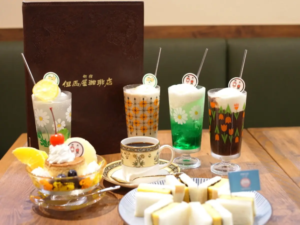 Assorted beverages in tall decorated glasses with straws and foam placed on a wooden table beside a pastry cake and white dessert slices