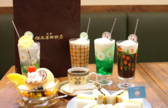 Assorted beverages in tall decorated glasses with straws and foam, placed on a wooden table beside a pastry cake and white dessert slices.