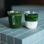 Two clear plastic cups hold matcha drinks on a blue tiled counter one fully green and the other layered with milk on the bottom