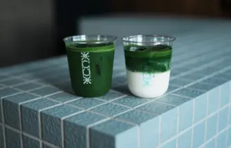 Two clear plastic cups hold matcha drinks on a blue tiled counter one fully green and the other layered with milk on the bottom