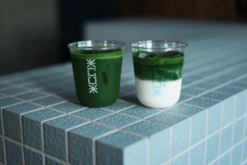 Two clear plastic cups hold matcha drinks on a blue tiled counter one fully green and the other layered with milk on the bottom