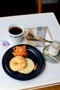 Plate with a bagel half topped with cream cheese and a side of shredded carrot salad on a dark blue plate with a cup of black coffee and a jar of yogurt nearby on a white table