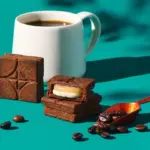 White mug of coffee beside stacked chocolate brownies on a teal surface with scattered coffee beans and an orange scoop nearby