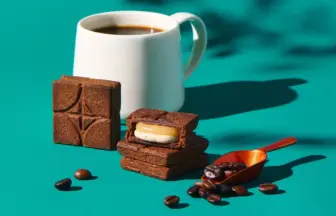 White mug of coffee beside stacked chocolate brownies on a teal surface with scattered coffee beans and an orange scoop nearby