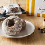 Powdered sugar donuts on a white plate with coffee beans nearby and a yellow and white Coffee Donut box in the background