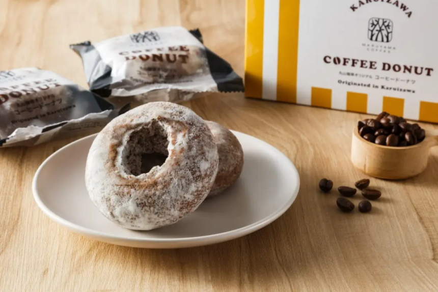 Powdered sugar donuts on a white plate with coffee beans nearby and a yellow and white Coffee Donut box in the background