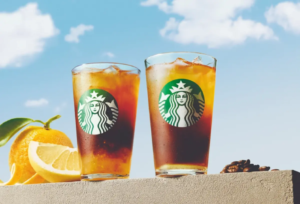 Two tall iced Starbucks drinks on a ledge with orange slices nearby against a bright blue sky