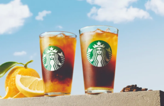 Two tall iced Starbucks drinks on a ledge with orange slices nearby, against a bright blue sky.