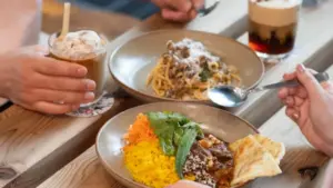 Two diners share a wooden table with plates of food a bowl of yellow rice with beef and greens plus naan and a pasta dish with two drinks nearby and hands reaching in to eat