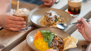 Three bowls of food on a wooden table with drinks nearby including a yellow rice bowl a pasta dish and a meat topped bowl as people reach for utensils and glasses of beverages nearby