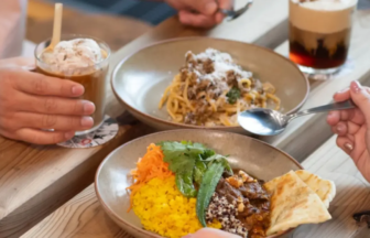 Three bowls of food on a wooden table with drinks nearby, including a yellow rice bowl, a pasta dish, and a meat-topped bowl, as people reach for utensils and glasses of beverages nearby.
