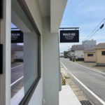 Street view showing a Marine Coffee sign hanging over a doorway along a quiet road with a sidewalk and blue sky above