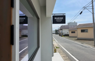 Street view showing a Marine Coffee sign hanging over a doorway along a quiet road with a sidewalk and blue sky above