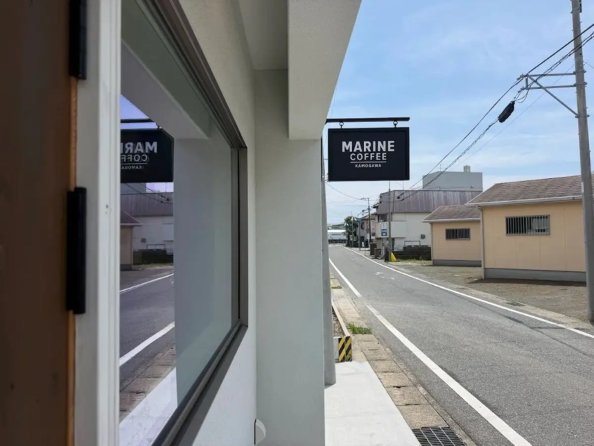 Street view showing a Marine Coffee sign hanging over a doorway along a quiet road with a sidewalk and blue sky above