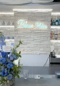 Neon sign reading Blue Miu on a white brick wall behind a cafe counter with a blue floral arrangement in the foreground