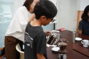 Boy with glasses pours hot water from a kettle into a pour over coffee setup on a table as adults watch nearby