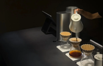 Pour over coffee station with three glass carafes paper filters and a hand pouring water from a kettle on a dark counter