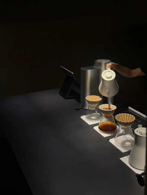 Pour over coffee station with three glass carafes paper filters and a hand pouring water from a kettle on a dark counter