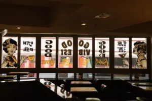 Dim restaurant interior with large front windows showing retro cartoon portraits and bold reversed text on glass